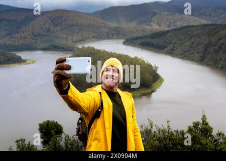 Sorridente in una giacca gialla che scatta un selfie con un telefono sullo sfondo di un tranquillo fiume circondato da paesaggi invernali. Foto Stock