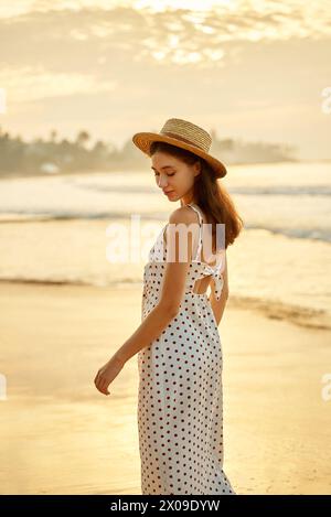 Elegante donna in abito a pois gode di un tramonto sulla spiaggia tropicale. La rilassata viaggiatrice femminile vaga a piedi nudi nella sabbia, mentre l'abito estivo scorre. Solo Foto Stock