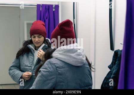 Donna di mezza età che prova una giacca e un cappello in un camerino in un punto di prelievo. La donna sta esaminando attentamente gli oggetti nello specchio per creare un Foto Stock