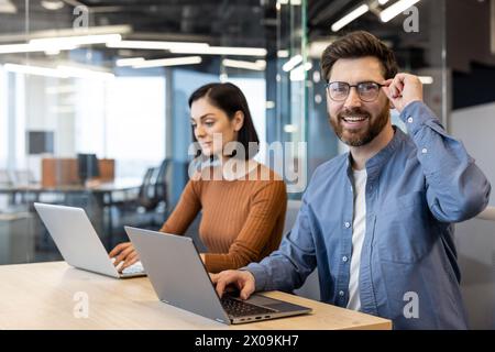 Un uomo e una donna lavorano fianco a fianco in un ufficio luminoso e moderno, utilizzando computer portatili e mostrando un atteggiamento positivo e mirato. Foto Stock