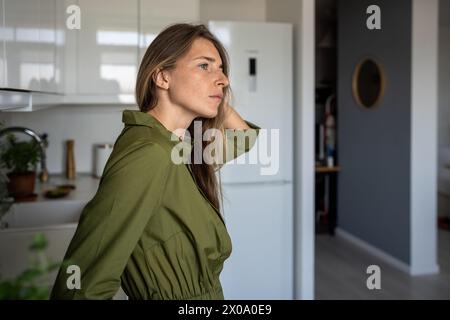 Una donna di mezza età, seria e premurosa, che purtroppo guarda, ha un cattivo umore in cucina a casa. Foto Stock