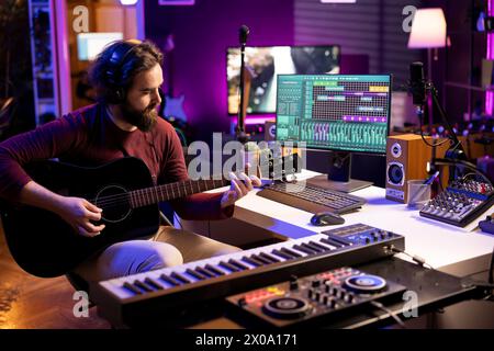 Giovane cantante che suona la chitarra acustica e crea musica con strumenti elettronici in casa, utilizzando l'interfaccia software daw su pc. Il produttore del suono registra i brani e mixa i suoni sulla console stereo. Foto Stock