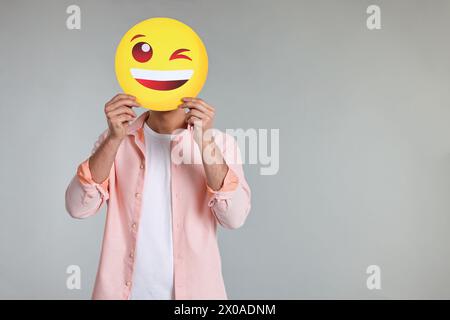 Uomo che ricopre il viso con emoticon allegro su sfondo grigio. Spazio per il testo Foto Stock