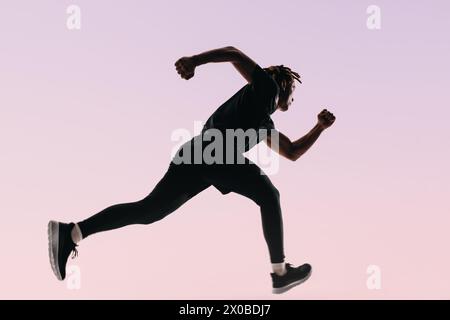 Atleta con dreadlock mette in mostra atletismo e forma in un allenamento ad alta intensità. Su uno sfondo rosa, la silhouette di un runner salta a mezz'aria, e. Foto Stock