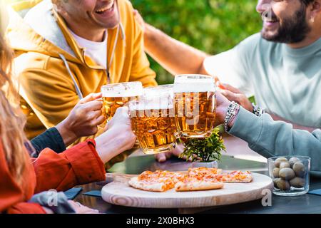 Un gruppo di amici tifa con tazze da birra su un tavolo rustico carico di pizza, gustando una festa conviviale in una birreria all'aperto. Foto Stock