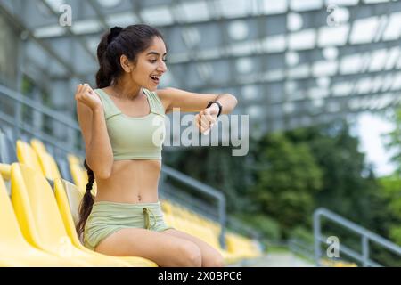 La giovane donna indiana allegra e sfidante siede sulla tribuna dello stadio e si rallegra del risultato, guarda l'orologio intelligente e il tracker, mostra un gesto di vittoria con la mano. Foto Stock