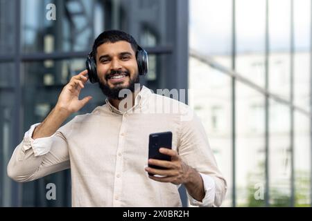 Ritratto ravvicinato di un giovane musulmano sorridente in piedi all'esterno con le cuffie, tenendo un telefono e ascoltando sorridendo davanti alla fotocamera. Foto Stock
