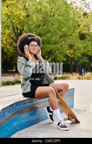 Una giovane donna afroamericana con i capelli ricci si siede su una panchina, coinvolta in una conversazione sul suo cellulare in uno skate Park. Foto Stock