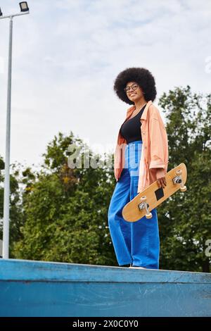 La giovane donna afroamericana con i capelli ricci tiene con sicurezza lo skateboard, adagiata sulla rampa dello skate Park. Foto Stock