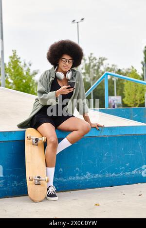 Una giovane donna afroamericana siede sul suo skateboard, vicino a una rampa in uno skate Park all'aperto. Foto Stock