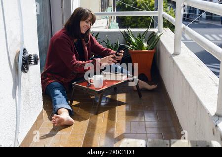 Donna latina adulta di etnia argentina seduta sulla terrazza del suo appartamento al mattino di sole, intrattenuta leggendo messaggi sul suo telefono mentre era rea Foto Stock