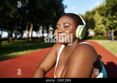 Una donna afroamericana in abbigliamento sportivo siede, indossa le cuffie, immersa nella musica, incarna la positività del corpo mentre è all'aperto. Foto Stock