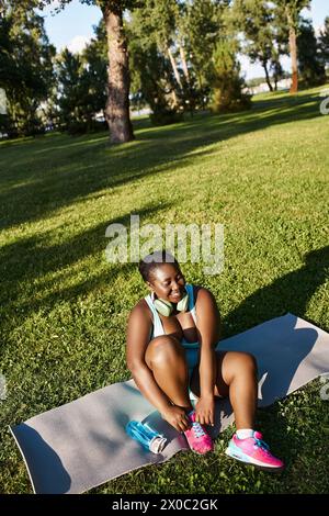 Una donna afroamericana in abbigliamento sportivo si siede su un tappeto all'aperto, con le cuffie Foto Stock