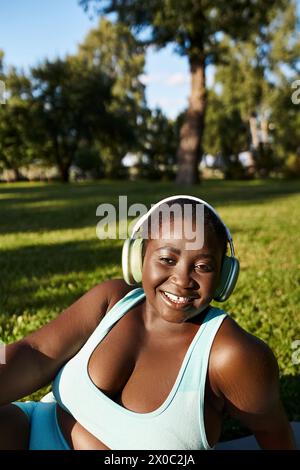 Una donna afroamericana in abbigliamento sportivo si siede sull'erba, ascoltando musica attraverso le cuffie, godendosi la vita all'aria aperta. Foto Stock