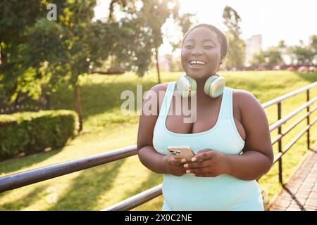 Una donna afroamericana sicura di sé, che indossa le cuffie, ama ascoltare musica mentre tiene in mano un cellulare all'aperto. Foto Stock