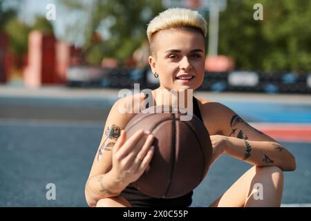 Una giovane donna con i capelli corti e i tatuaggi si siede per terra, cullando un pallacanestro con un look deciso. Foto Stock