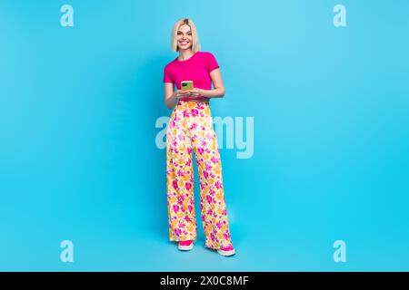 Foto a tutto il corpo di una giovane donna attraente tenere il telefono eshop vestiti eleganti rosa isolati su sfondo blu Foto Stock