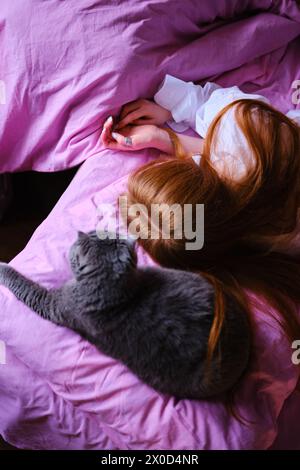 bella giovane donna allo zenzero con il suo gatto britannico grigio nel letto rosa a casa ragazza in camicia bianca Foto Stock