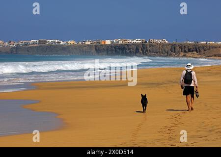 Un uomo solitario e il suo cane camminano lungo una spiaggia sabbiosa lasciando impronte, El Cotillo, Fuerteventura. Preso nel febbraio 2024 Foto Stock