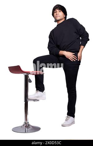 Un giovane latino adulto sta posando usando una sedia da parrucchiere. Il giovane è vestito di nero e lo sfondo è bianco. Foto Stock