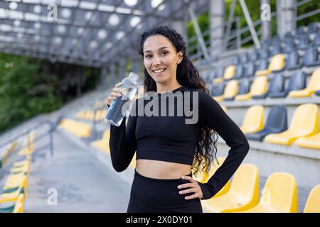 Una donna ispanica in forma che fa una pausa per bere acqua durante la sua routine di esercizio in uno stadio vuoto. Foto Stock