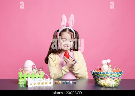 Adorabile bambina che colorava le uova con acquerello e pennello per preparazioni pasquali, decorando ornamenti natalizi su sfondo rosa. I bambini piccoli si divertono con le decorazioni. Foto Stock