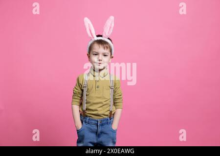 Giovane e gioioso bambino che indossa simpatiche orecchie da coniglietto davanti alla macchina fotografica, si sente entusiasta della festa di pasqua e in piedi su sfondo rosa. Piccolo allegro con un vestito adorabile. Foto Stock