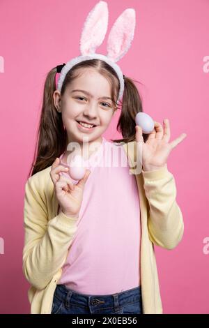 Giovane ragazza carina che posa con sicurezza davanti alla macchina fotografica, presentando le sue uova rosa dipinte per la festa di pasqua. Una piccola studentessa che indossa orecchie da coniglio sorridente in studio, mostra decorazioni fatte a mano. Foto Stock