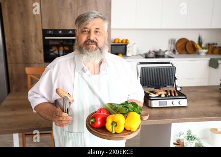 Uomo maturo con tavola di verdure fresche e gustosa salsiccia cucinata su un moderno grill elettrico al tavolo della cucina Foto Stock