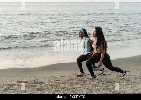 Le donne in allenamento eseguono affondamenti su una spiaggia sabbiosa con dolci onde sullo sfondo. Foto Stock