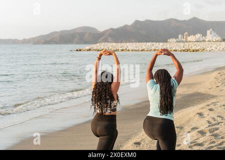 Due donne si allungano di fronte al mare, con le loro silhouette contro la luce del tramonto, con montagne e paesaggi urbani alle spalle. Foto Stock