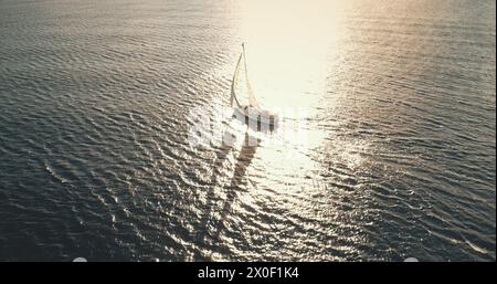 Riflesso del sole sullo yacht che naviga sull'antenna della baia cristallina del mare. Mesmerezing Seacape con barca a vela in estate, giorno di luce solare intensa. Tranquillo lungomare con barca a vela da sola al soffio del vento al colpo del drone Foto Stock