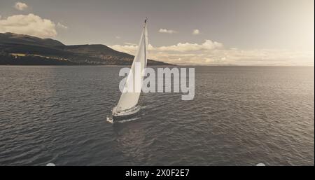 Riflesso del sole sullo yacht che naviga sull'antenna della baia cristallina del mare. Mesmerezing Seacape con barca a vela in estate, giorno di luce solare intensa. Tranquillo lungomare con barca a vela da sola al soffio del vento al colpo del drone Foto Stock