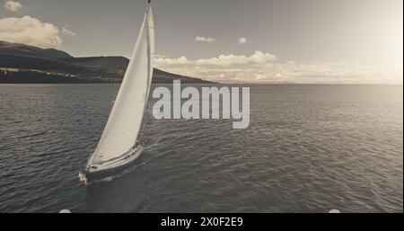 Riflesso del sole sullo yacht che naviga sull'antenna della baia cristallina del mare. Mesmerezing Seacape con barca a vela in estate, giorno di luce solare intensa. Tranquillo lungomare con barca a vela da sola al soffio del vento al colpo del drone Foto Stock