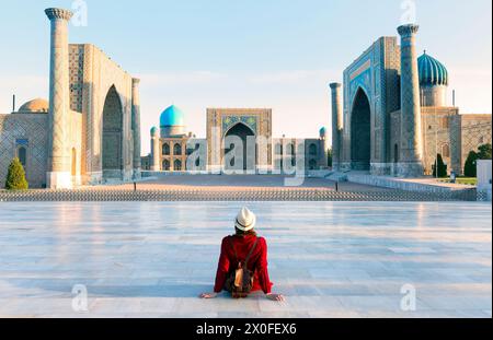 Donna turistica con cappello e abito rosso seduta su Registan, un'antica piazza pubblica nel cuore dell'antica città di Samarcanda, Uzbekistan. Foto Stock