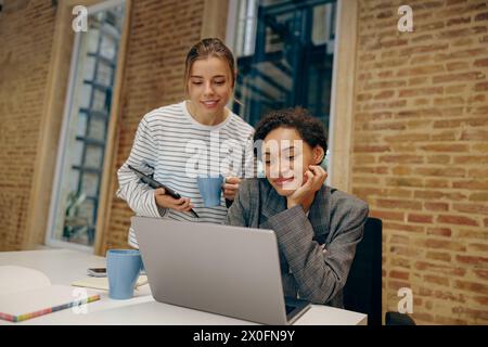 Due colleghi donne concentrate che lavorano insieme al progetto e utilizzano un laptop seduto in ufficio Foto Stock