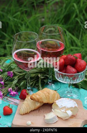Colazione romantica in giardino. Fine settimana del giorno d'estate. Due bicchieri di spumante, croissant, fragole, formaggio e fiori rosa su un tavolo. Foto Stock