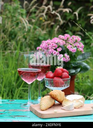Colazione romantica in giardino. Fine settimana del giorno d'estate. Due bicchieri di spumante, croissant, fragole, formaggio e fiori rosa su un tavolo. Foto Stock