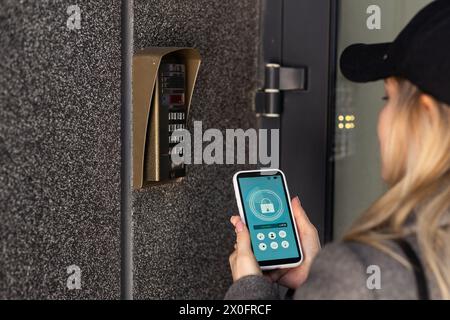 Donna che blocca lo smartlock sulla porta d'ingresso utilizzando uno smartphone. Concetto di utilizzo di serrature elettroniche intelligenti con accesso senza chiavi. Foto Stock