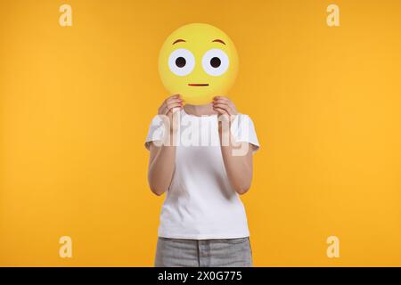 Donna che copre il viso con emoticon sorpresa su sfondo giallo Foto Stock