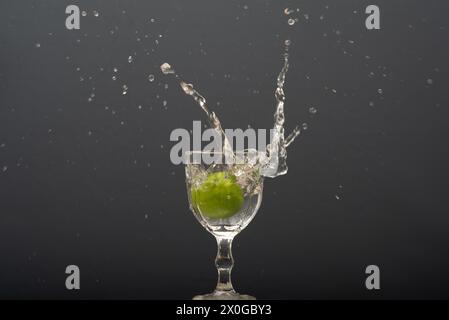 Il limone cade in un bicchiere d'acqua trasparente causando schizzi verso l'alto. Foto Stock