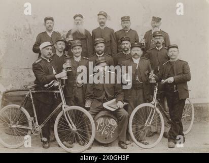 Un gruppo di 14 servi nella loro posa uniforme per una foto. Indossano tappi e tengono in mano una tazza di birra. Il segretario è seduto per una birra. I due uomini sul lato tengono le loro biciclette. Uno degli uomini sta fumando, molti stanno trasportando gli orologi della tasca. La data del giorno della foto è scritta sulla tazza della birra. [traduzione automatica] Foto Stock