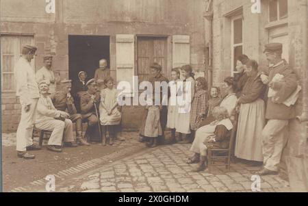 Soldati in uniforme bianca e grigia da campo siedono e si ergono in un cortile con ciottoli grezzi di fronte a un grande gruppo di locali francesi. Stanno chiacchierando e ridendo. I francesi sono casuali, così come i soldati tedeschi. Un soldato, probabilmente un ufficiale, si appoggia alla sua sciabola [traduzione automatica] Foto Stock