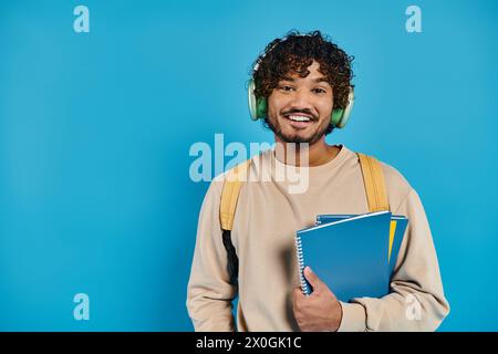 felice uomo indiano che indossa le cuffie, tiene i libri e sorride su sfondo blu Foto Stock