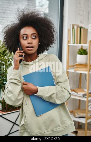 una donna nera di diversa provenienza parla al telefono cellulare mentre tiene una cartella in un moderno spazio di coworking. Foto Stock