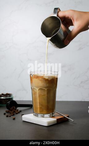 Versare il latte d'avena in un caffè freddo ghiacciato in un alto tavolo di vetro in marmo grigio Foto Stock