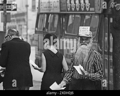 Ein älterer Mann mit langem Bart und einer Mütze mit der Aufschrift 'Christ Saves Sinners' verteilt christliche Botschaften an Passanten an einer Straße a Manhattan, New York City. (Aufnahmedatum: 01.01.1967-31.12.1967) Foto Stock