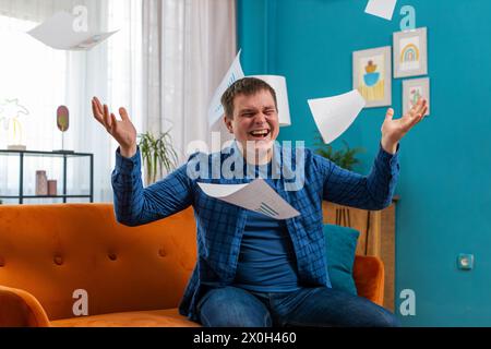 Stressato giovane freelance caucasico che esamina grafici e grafici aziendali e lancia in aria carte non pagate, seduti sul divano nel salotto di casa. Un tizio arrabbiato lavora a distanza sul divano dell'appartamento Foto Stock