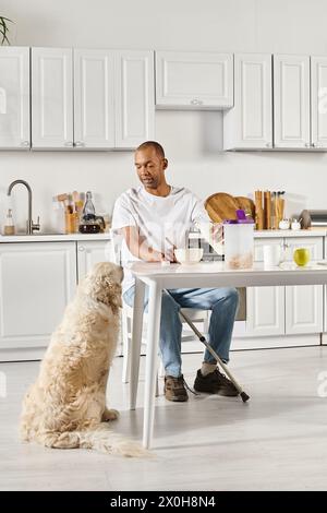 Un uomo afroamericano disabile siede a un tavolo da cucina con il suo leale cane Labrador al suo fianco. Foto Stock