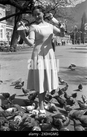 Una giovane donna in un vestito di colore chiaro che alimenta i piccioni in un parco. [traduzione automatica] Foto Stock